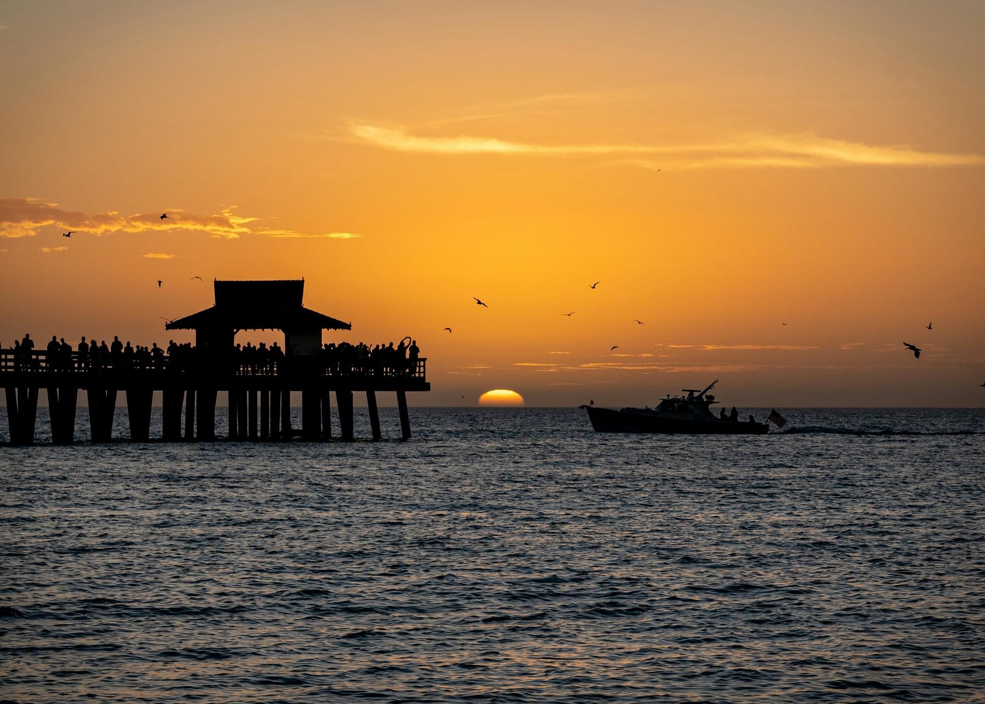 Silhouette of a pier with people and a boat at sunset over the ocean, with birds flying in the sky.