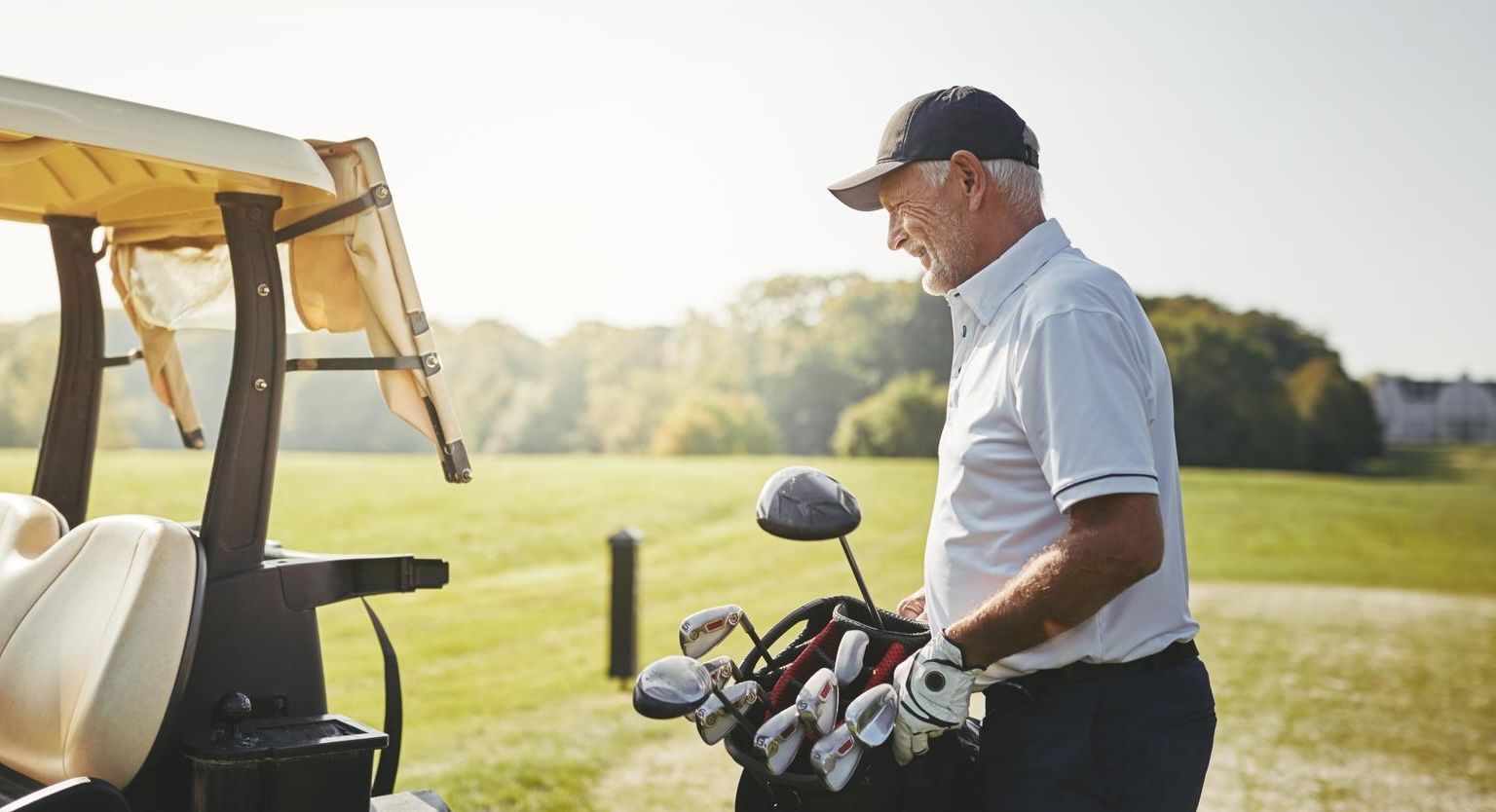 Older man in golf attire stands by a golf cart, smiling while holding a golf bag on a sunny course.