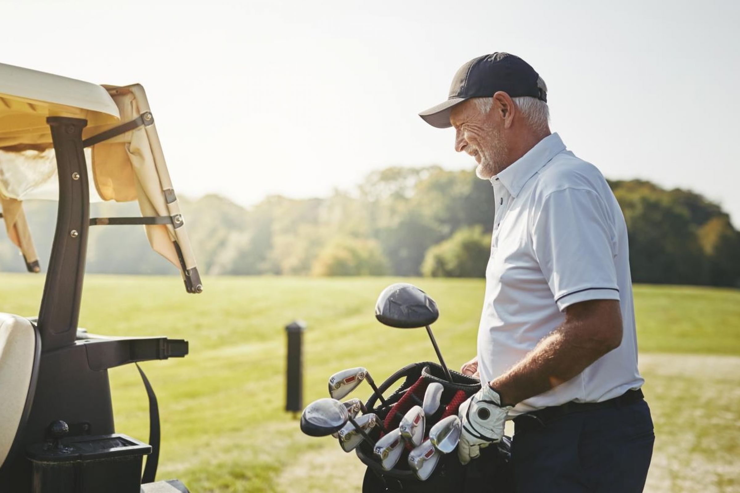 Older man in golf attire stands by a golf cart, smiling while holding a golf bag on a sunny course.
