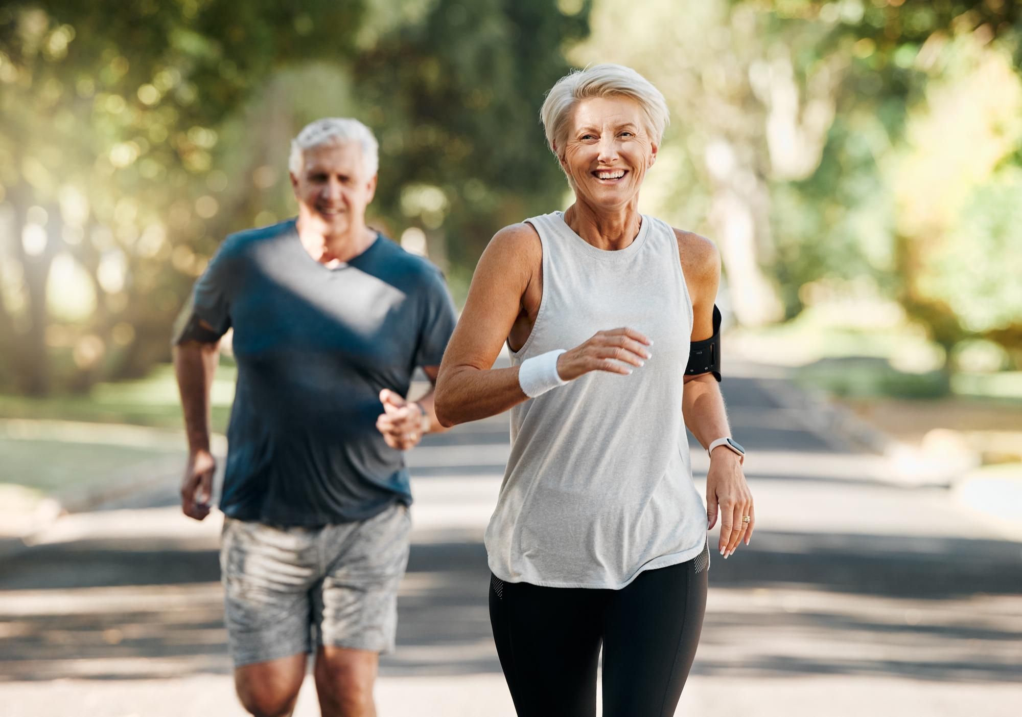 Smiling older woman jogging outdoors, with an older man running behind her on a sunny day.
