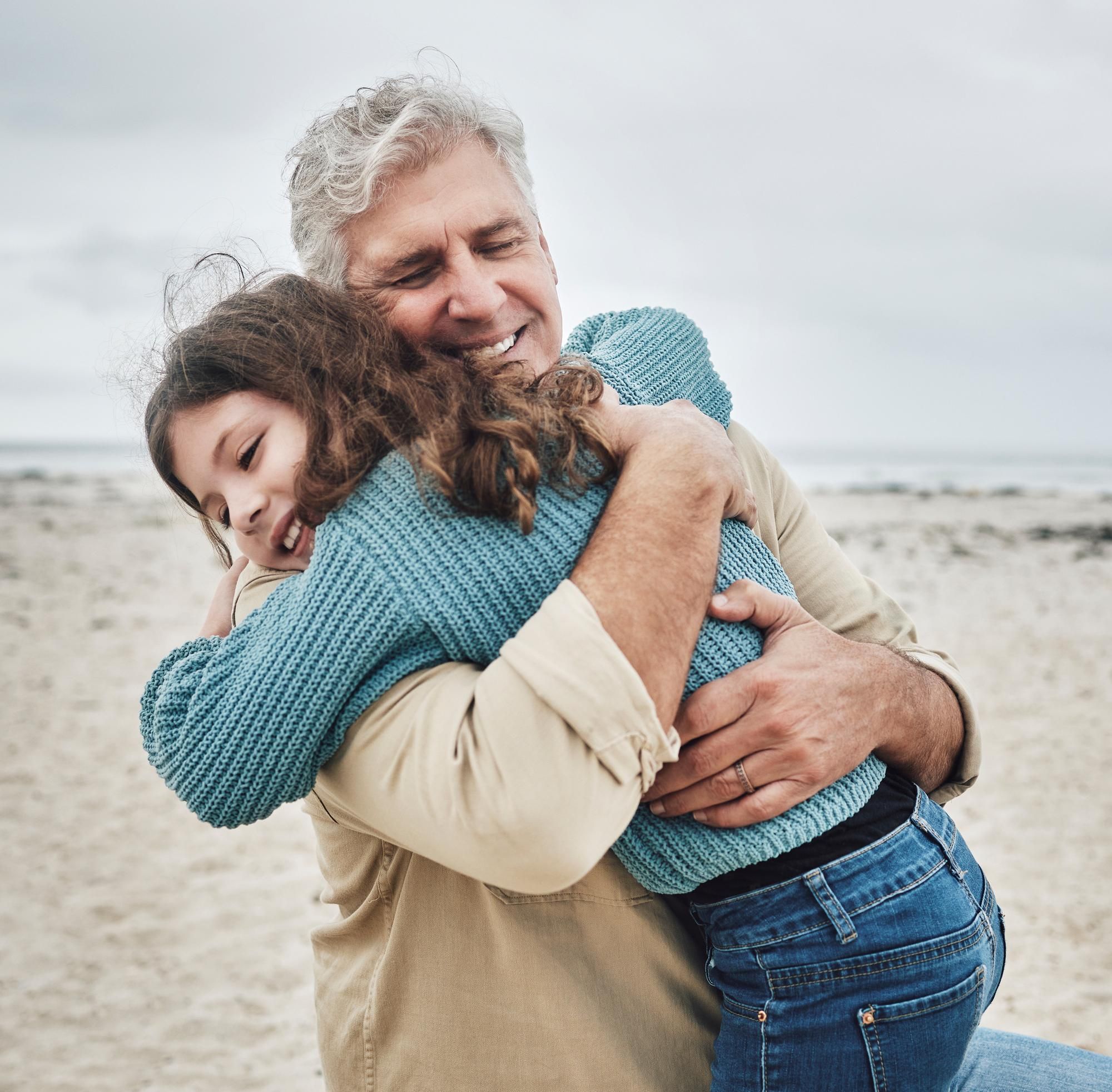Older man hugging a young girl on a beach, both smiling with eyes closed, enjoying a warm embrace.