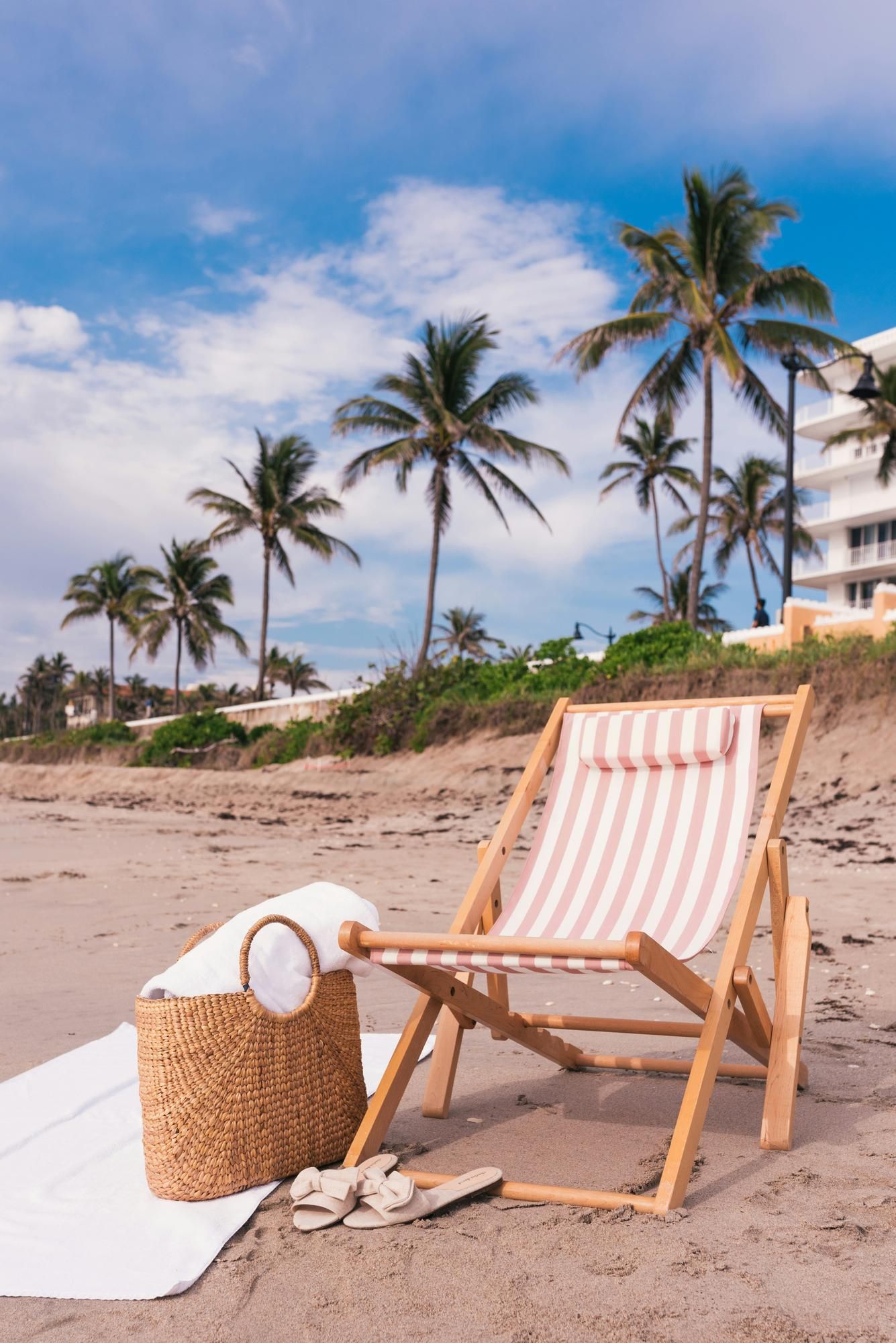 A striped beach chair, towel, basket, and sandals on sandy beach with palm trees and blue sky.